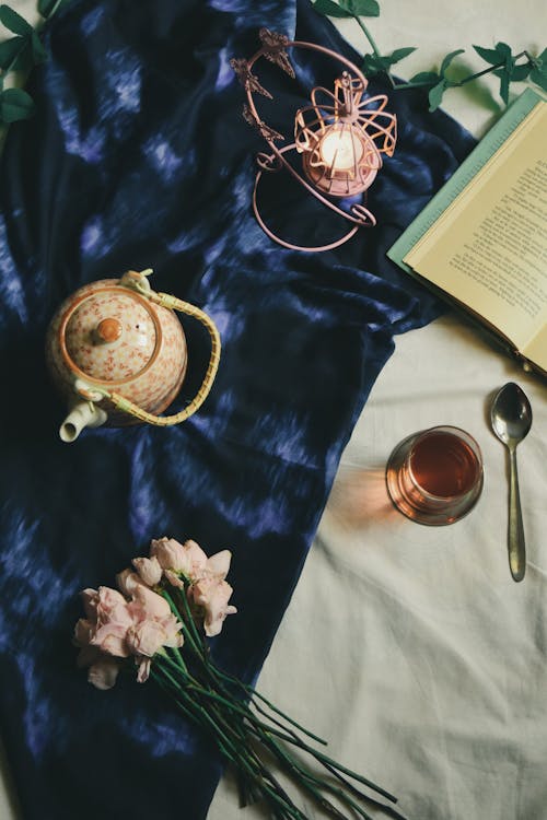 Free From above arrangement of ceramic teapot and glass of hot fresh tea served on blue silk cover on bed near book flowers and candleholder Stock Photo