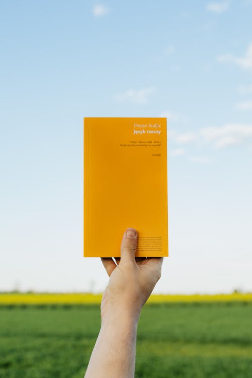 Crop anonymous person holding up orange book against clear blue sky on summer day in green field