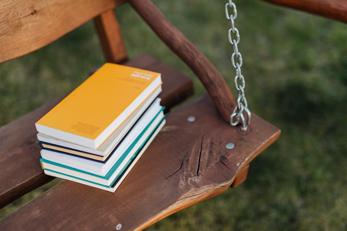 From above of pile of colorful books on wooden bench of garden swing on quiet summer evening