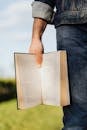 Crop unrecognizable male student in casual outfit standing with book in hand on green spring meadow with sunshine lighting up pages of novel