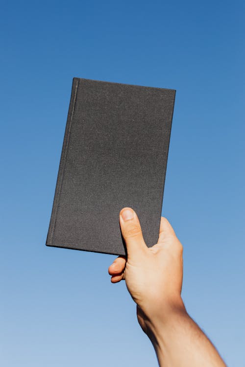 Free Crop unrecognizable person with closed black notebook in hand against clear blue sky Stock Photo