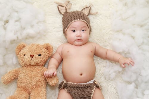 無料 ヒグマぬいぐるみの横にある白いクッションの上に横たわっている赤ちゃん 写真素材