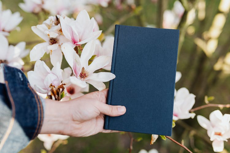 Closed Book In Hand Of Anonymous Person Against Flower Background On Sunny Day