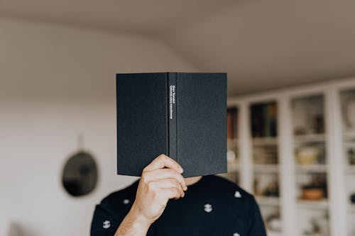 Faceless male holding opened black book in hand covering face while standing in middle of light room