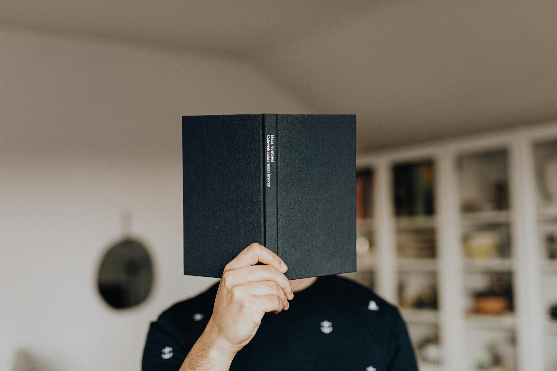 Faceless male holding opened black book in hand covering face while standing in middle of light room