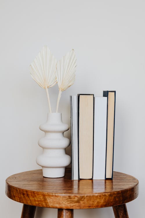 White original vase on wooden round table along with stack of assorted books in front of white wall