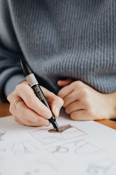 Crop anonymous female artist in casual wear with ring on finger using marker while drawing on paper with drafts at wooden table at work
