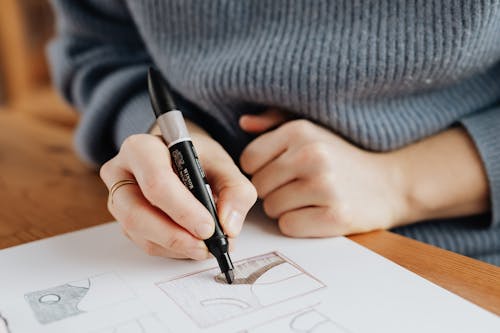 Crop unrecognizable female designer in warm sweater drawing with brown marker on large sheet of paper while working on pattern