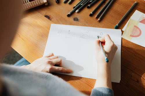 From above of crop faceless female designer in casual clothes creating scheme on sheet of paper with pencil while sitting at wooden desk in sunlight