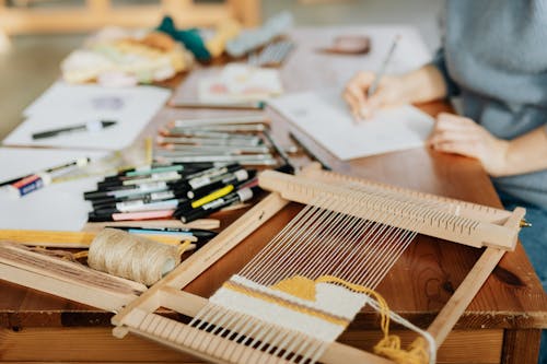 Crop unrecognizable female designer making sketch on paper while sitting at desk near various markers and pencils with wooden weaving loom machine with stretched threads