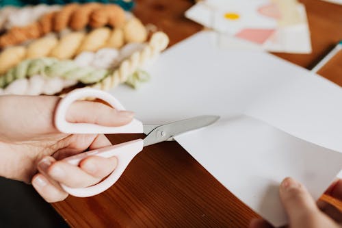 Crop young woman cutting paper sheet on wooden table