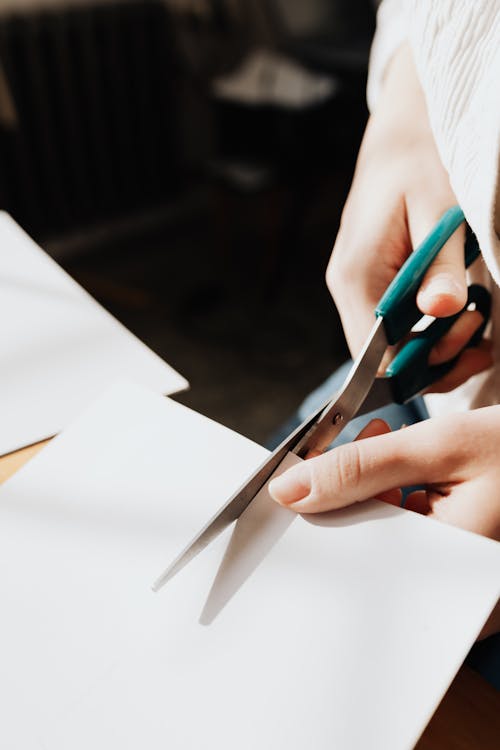 Unrecognizable female artisan cutting piece of paper with scissors during designing process on blurred background of workshop or atelier