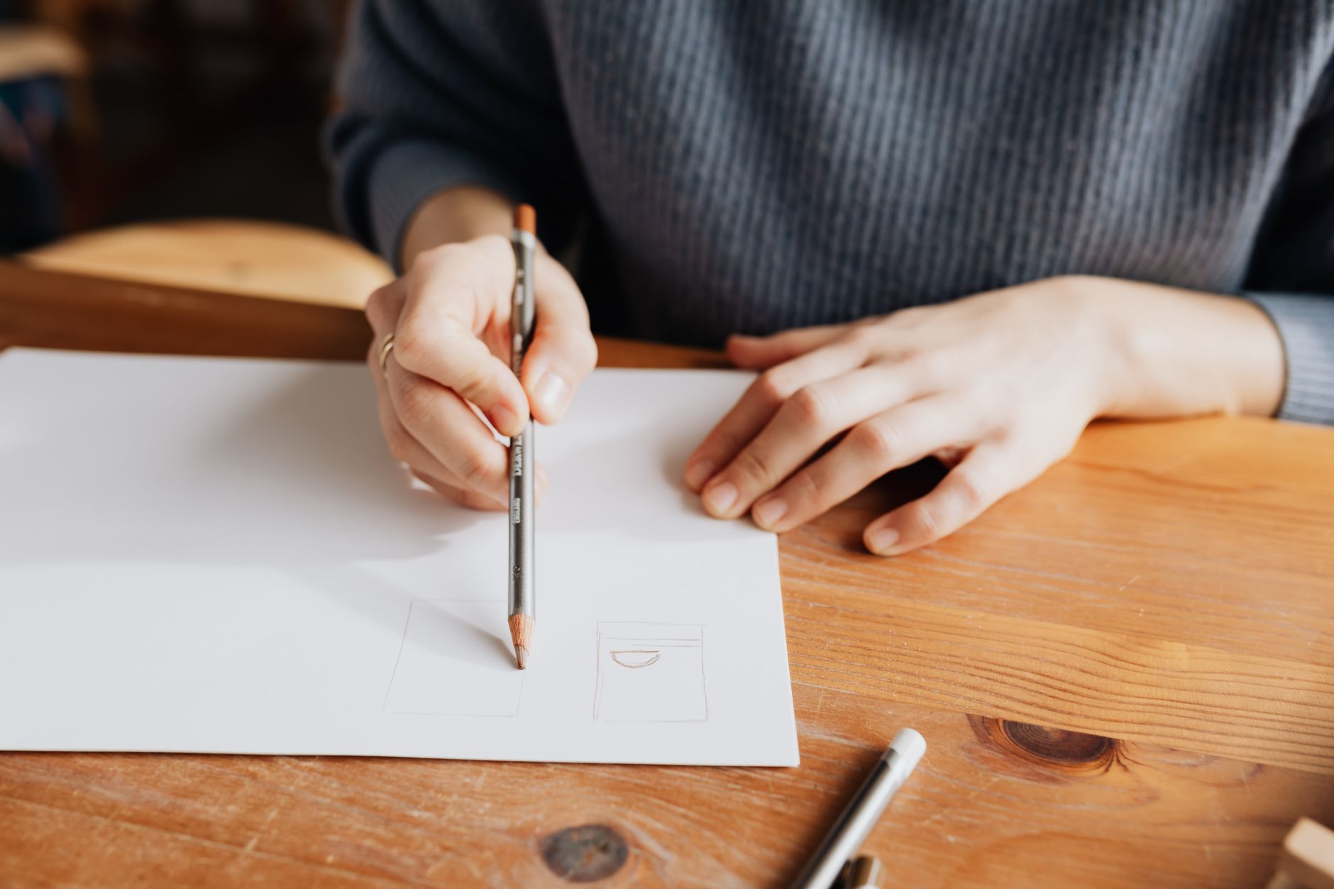 A person sketching on white paper with a pencil at a wooden table