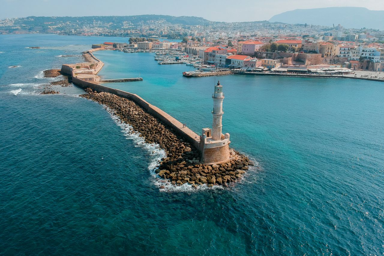 Drone capture of Chania's historic Venetian harbor, showcasing the iconic lighthouse against a scenic coastal backdrop.