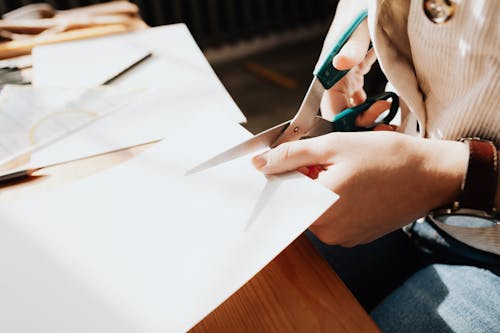 Unrecognizable young craftswoman cutting paper sheet on wooden table