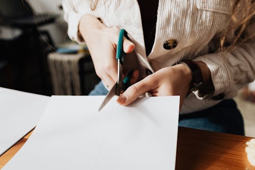 From above crop young craftswoman cutting piece of paper with scissors while sitting at wooden desk during designing process in workshop or atelier