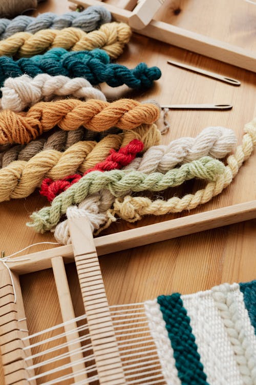 From above of unfinished tapestry on manual wooden loom placed on wooden table with assorted yarn twists and wooden needles during weaving process in workshop