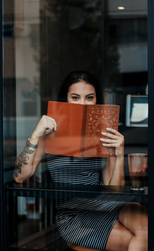 Through glass of unrecognizable positive young tattooed female resting in cafe and covering mouth with menu