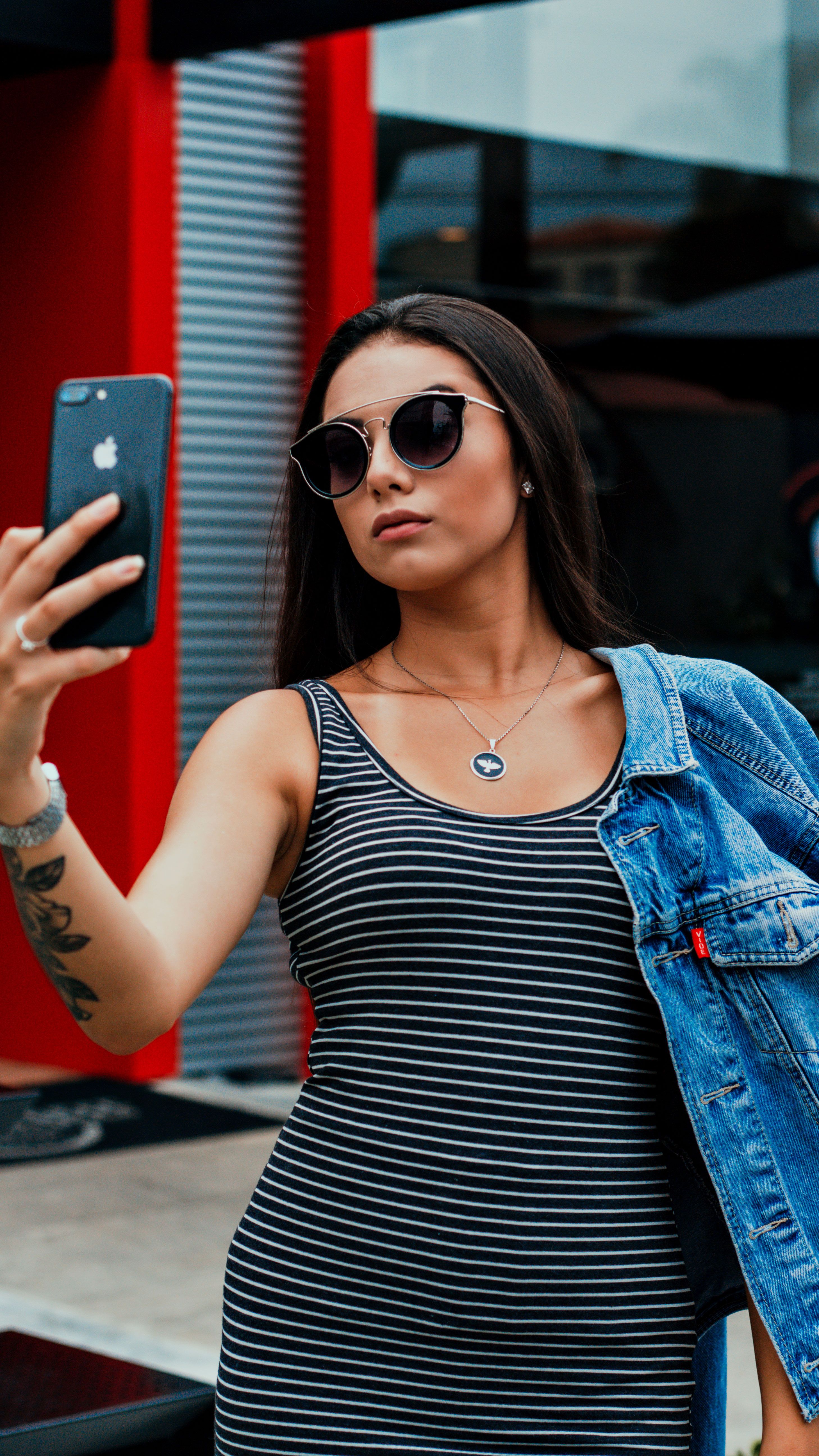 Self assured young tattooed woman in casual clothes and sunglasses taking selfie on smartphone while standing on street near modern building