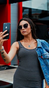 Self assured young tattooed woman in casual clothes and sunglasses taking selfie on smartphone while standing on street near modern building