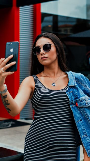 Self assured young tattooed woman in casual clothes and sunglasses taking selfie on smartphone while standing on street near modern building