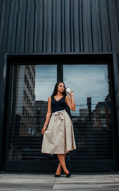 Full body of stylish young female with long dark hair in elegant outfit standing near entrance of modern building and drinking takeaway coffee