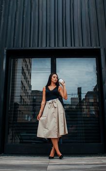 一位身着优雅服装、留着深色长发的时尚年轻女性，正站在现代建筑入口附近，喝着外卖咖啡