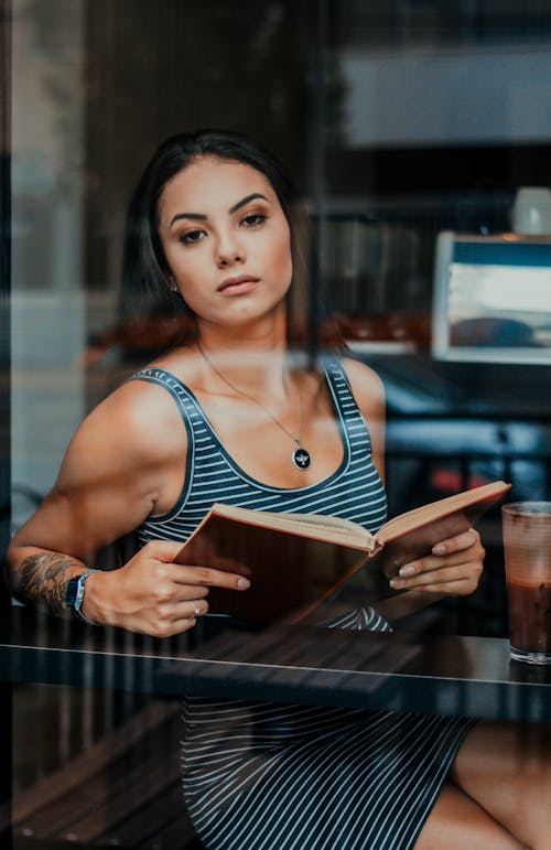 Through glass of focused young female with dark hair in casual outfit sitting in cafe with fresh beverage near window and reading interesting book