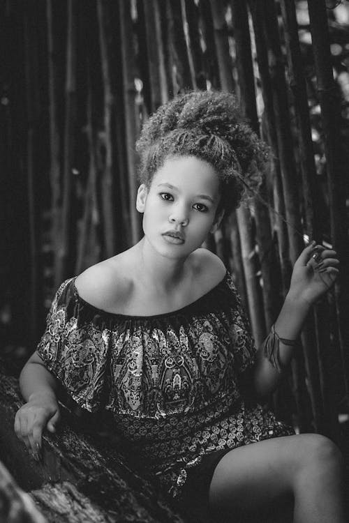 Black and white of serious young African American female with curly hair in stylish dress relaxing in tropical park among bamboo trees