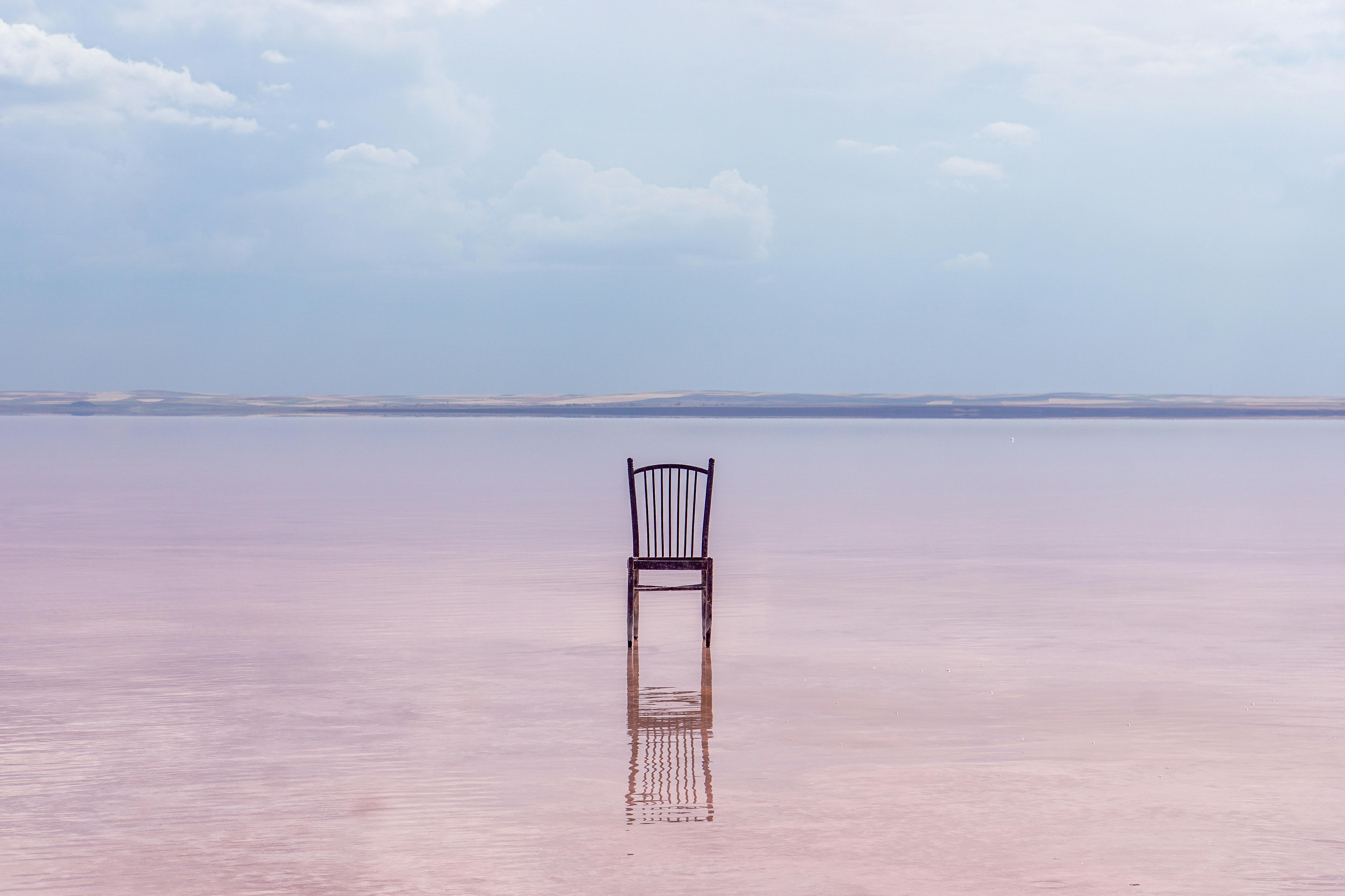 A wooden chair stands alone on a serene pink lake, reflecting the calm sky above.