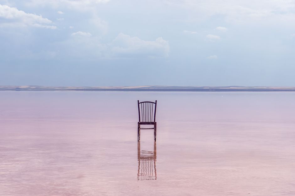 A wooden chair stands alone on a serene pink lake, reflecting the calm sky above.