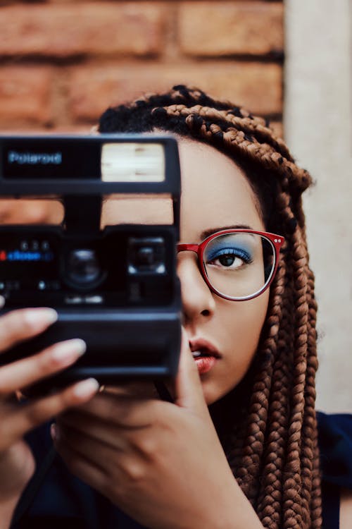 Focused young ethnic lady with Afro braids in stylish eyeglasses taking picture with vintage instant photo camera against brick wall