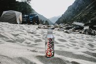 Coors Light bottle on a sandy campground with mountains and tents in the background.