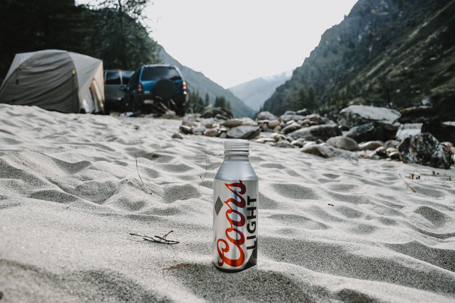 Coors Light bottle on a sandy campground with mountains and tents in the background.