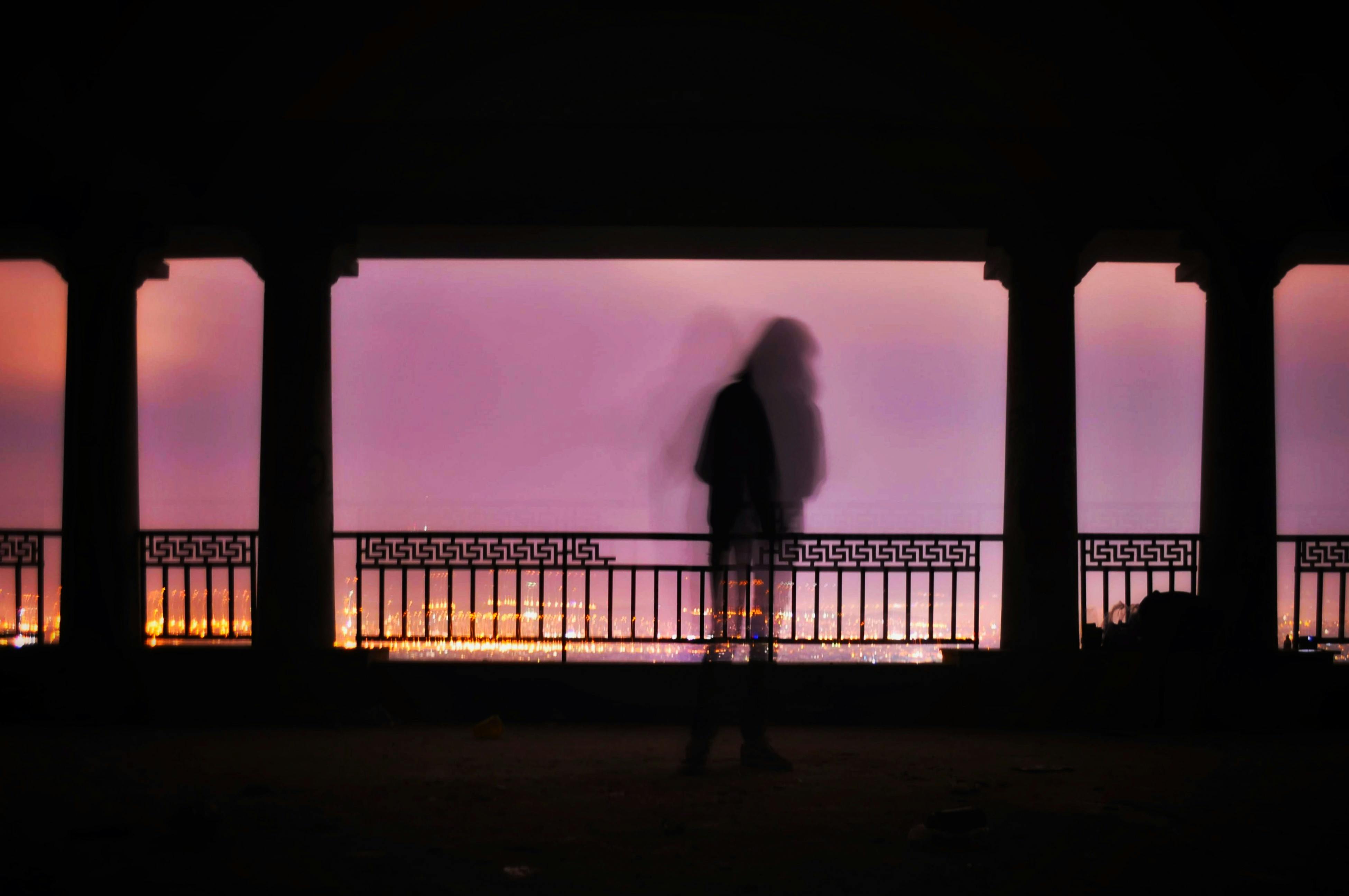 Silhouette of unrecognizable person standing on terrace of aged columned building against picturesque purple sunset sky over illuminated city