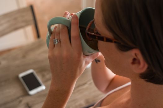 A woman with glasses drinks coffee from a mug indoors, smartphone on table.