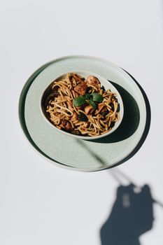 Overhead view of noodle dish garnished with herbs on a light plate casting shadows.