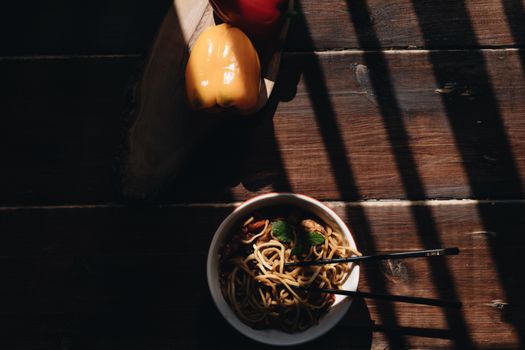 A bowl of Asian noodles with chopsticks, highlighted by shadows and sunlight, alongside bell peppers.