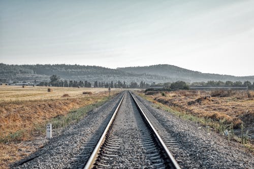 Straight railroad tracks going through dry grassy fields in countryside near mountains on sunny day