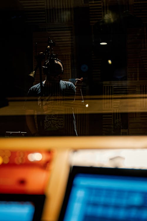 Through glass of musician in headphones recording song into microphone in modern music studio