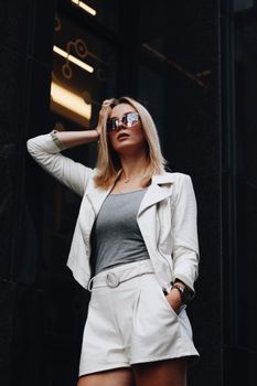 一位自信优雅的女性戴着时髦的太阳镜，手插在口袋里，靠在墙上