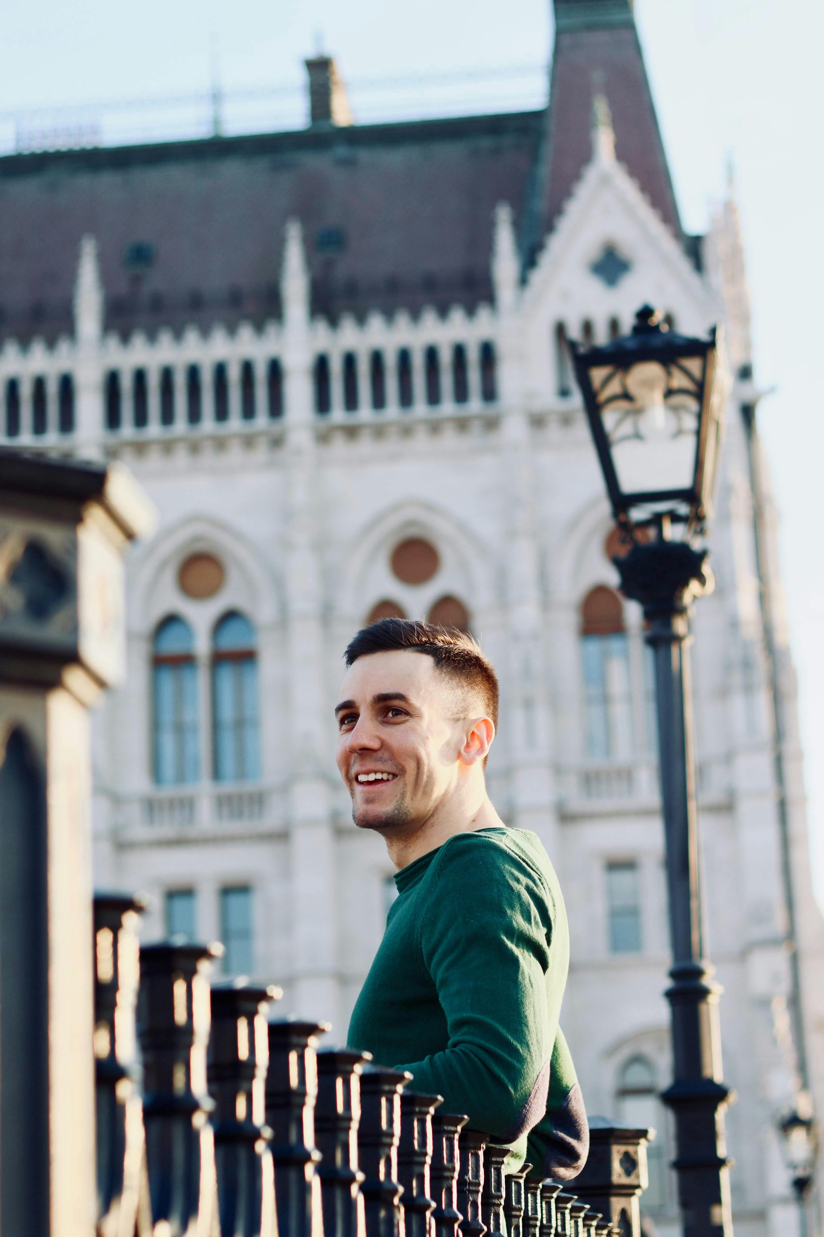 Low angle side view of cheerful young guy standing near metal fence and old building and looking away