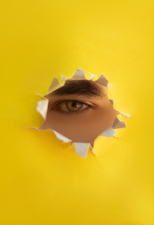 Free A close-up of an eye looking through a hole in vibrant yellow paper, creating a striking visual. Stock Photo