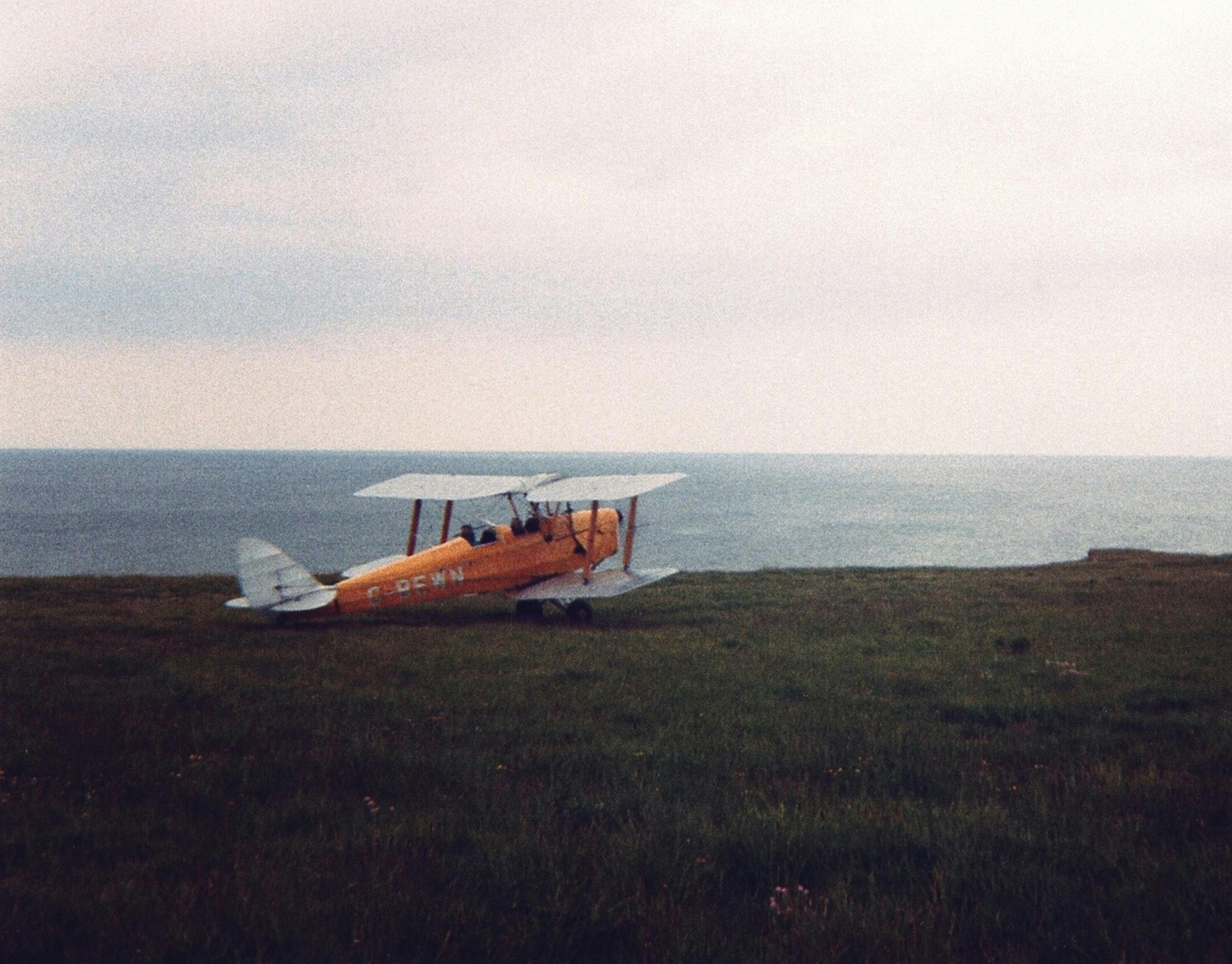 A classic yellow biplane rests on a grassy coastal landscape by the sea.