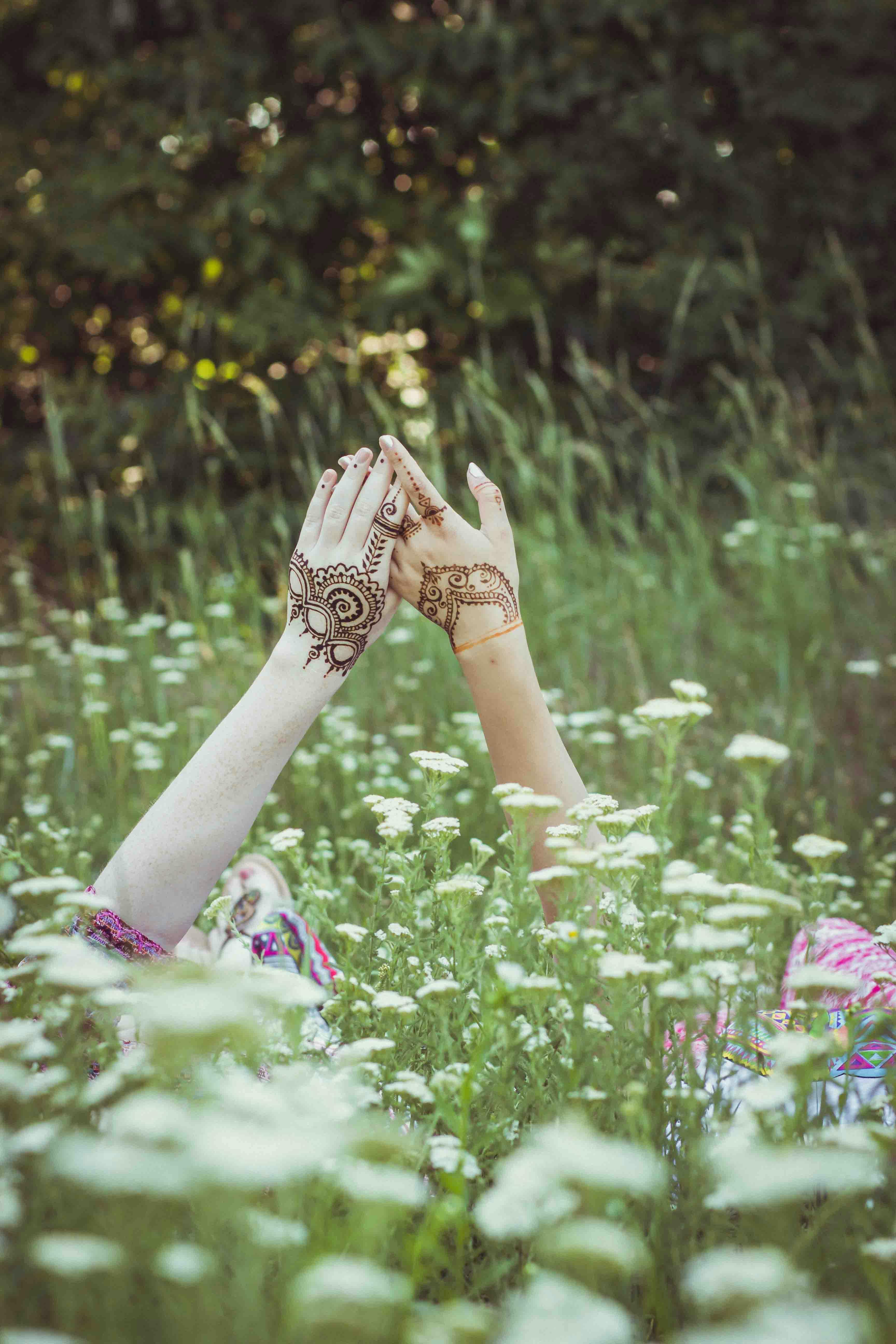 Hands with henna tattoos raised among wildflowers in a serene field setting.
