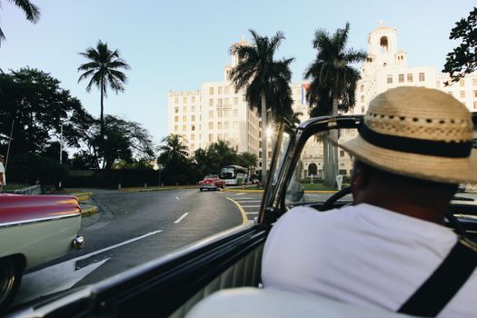Man driving a vintage convertible past the iconic Hotel Nacional de Cuba under palm trees.