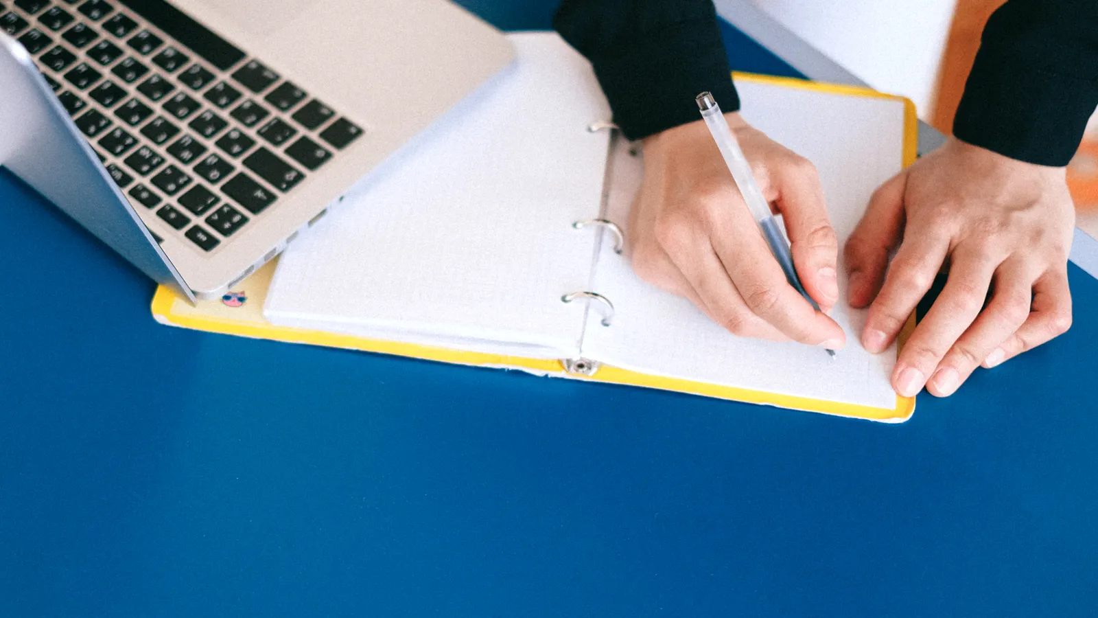 Hands writing notes in a notebook next to a laptop on a desk