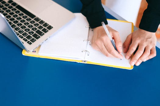Hands writing in a notebook with a laptop nearby on a blue table.