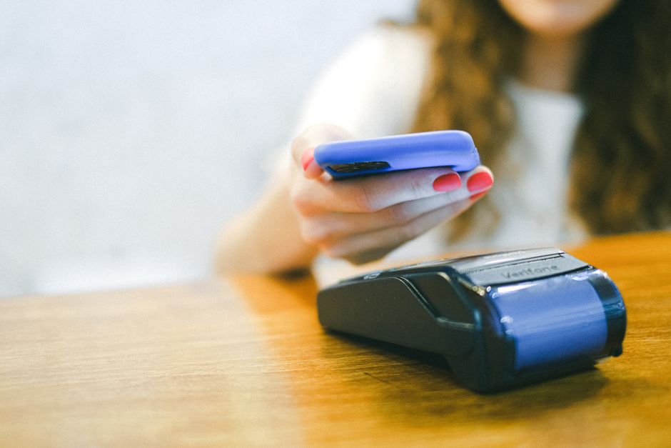 Dark-skinned Nigerian woman in a bright Ankara blouse using a smartphone to process a customer transaction at a food stall in a Lagos open market, other market stalls visible in the background, natural daylight, photorealistic