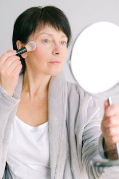 Elderly woman applying makeup using brush and mirror at home.